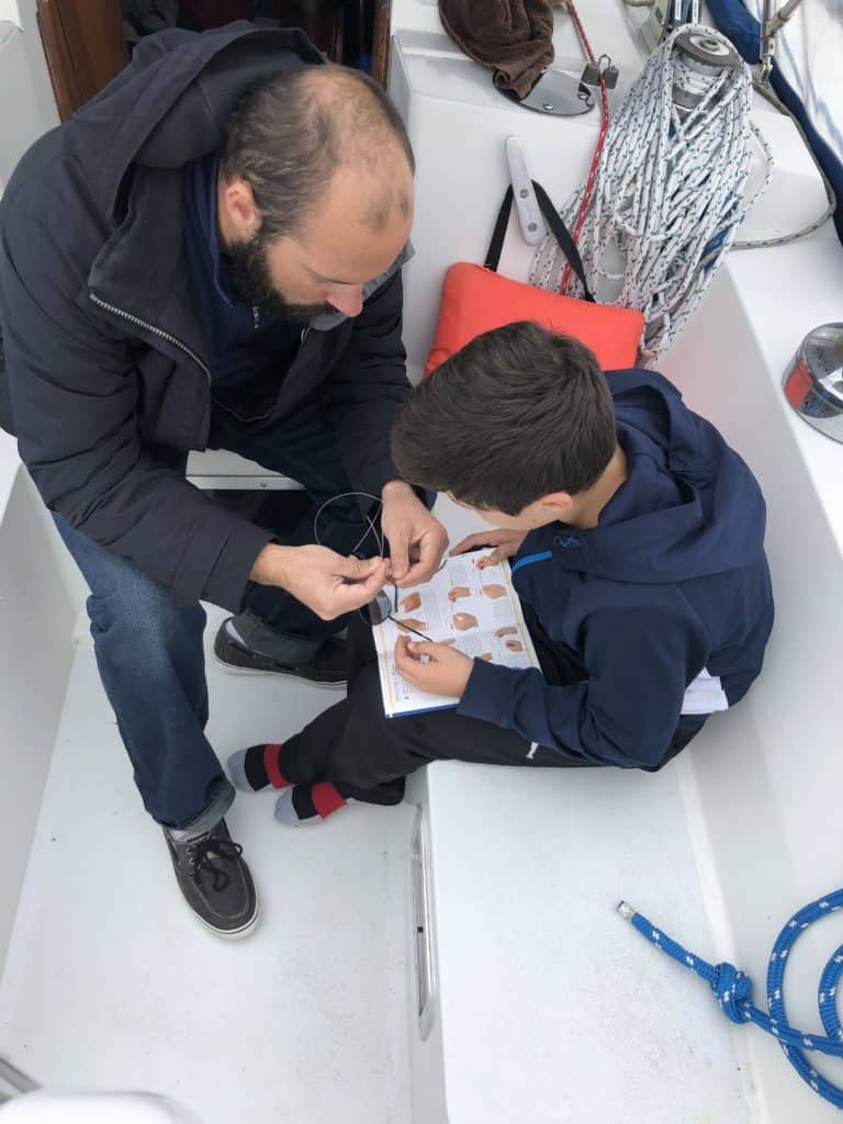 A father showing his son how to tie a knot, on a boat, using a knot book. Father is wearing Sperry sailing shoes. 