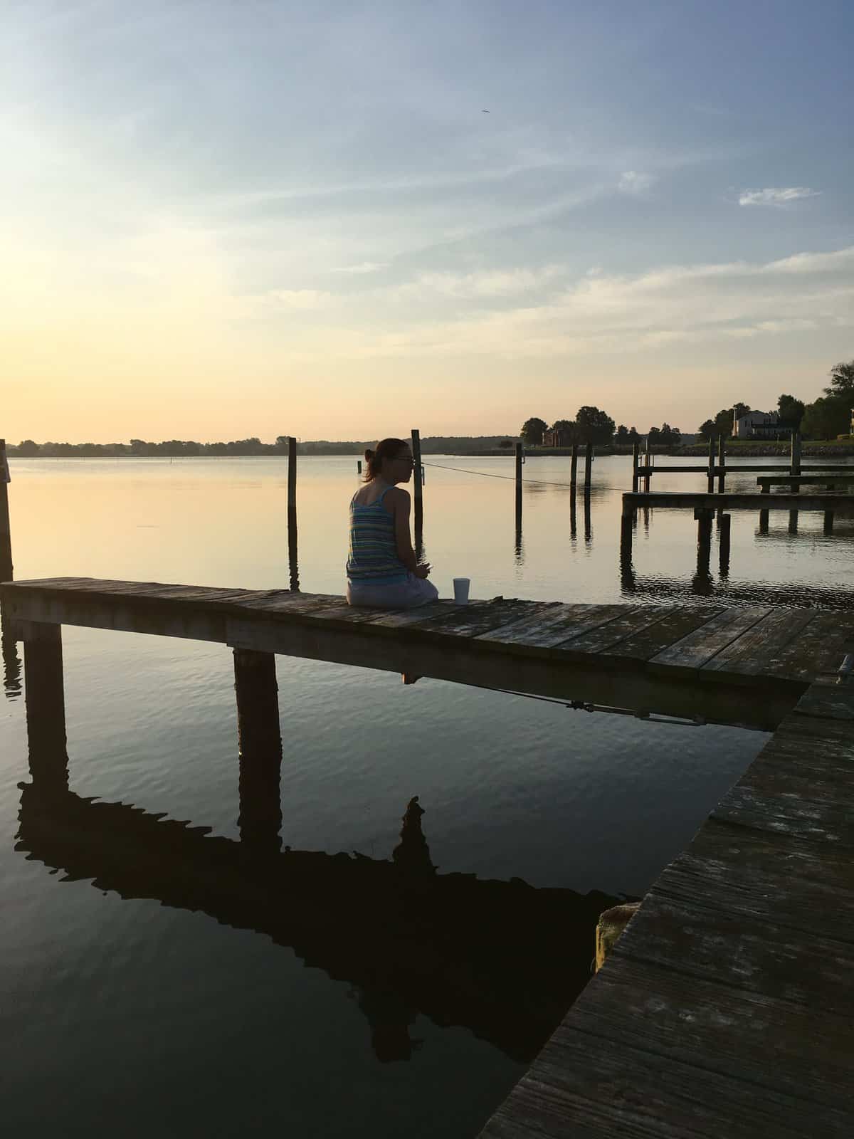 A woman sitting on the docks with a cup of coffee, during sunrise. 