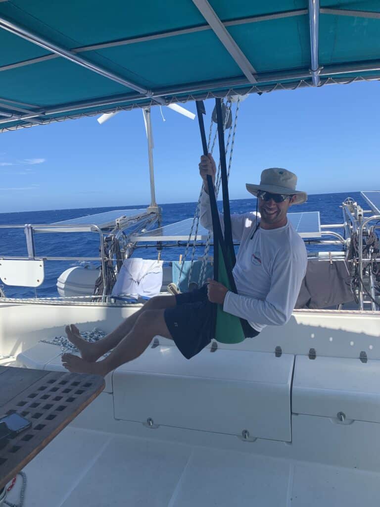 a man on a sailboat wearing a long sun shirt, short and a hat, swinging on a swing